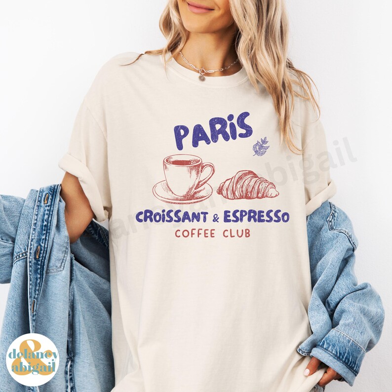 May include: A beige t-shirt with a graphic design that says "Paris Croissant & Espresso Coffee Club". The graphic includes a cup of coffee, a croissant, and a leaf.