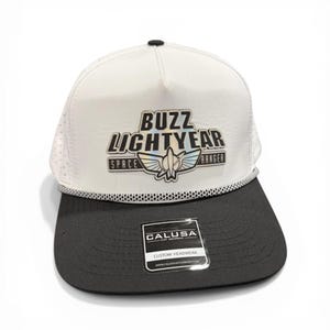 May include: White and black Buzz Lightyear Space Ranger hat. The hat features a white mesh top with the words "Buzz Lightyear Space Ranger" and a graphic of a spaceship. The brim is black, and the hat has a black and white rope detail.