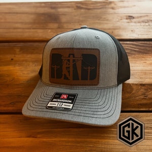 May include: Gray trucker hat with a black mesh back and a curved brim. The front features a brown leather patch with the word "DAD" and a silhouette of a lineman working on power lines. The hat has an adjustable strap and a Richardson tag.