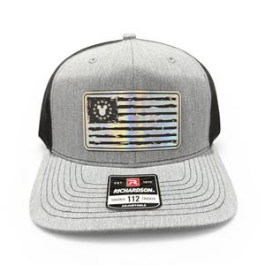 May include: Gray and black trucker hat with a patch featuring a distressed American flag design with a Mickey Mouse head in the stars. The hat has a curved brim and an adjustable strap. The patch reads "RICHARDSON ORIGINAL 112 TRUCKER ADJUSTABLE."