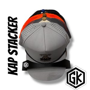 May include: A stack of baseball caps in various colors, including gray, orange, and black, displayed on a black stand. The front cap is gray with the text "KINDA FIT SORTA FAT". The image also features the text "KAP STACKER" and a "GK" logo.
