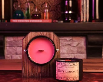 May include: A pink candle in a black and wooden holder with a black candle jar with a label that reads "Curio Constructs Fairy Garden" with a floral design. The candle jar is on a wooden surface.