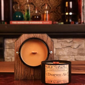 May include: A wooden framed candle with a circular candle and a black candle with a label that reads "Dwarven Ale". The candle is in a black container with a wooden wick. The background includes a shelf with bottles and a book.