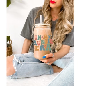 May include: A mason jar with a straw and a lid, filled with a light brown beverage. The jar has a colorful graphic that says "In My Auntie Era".