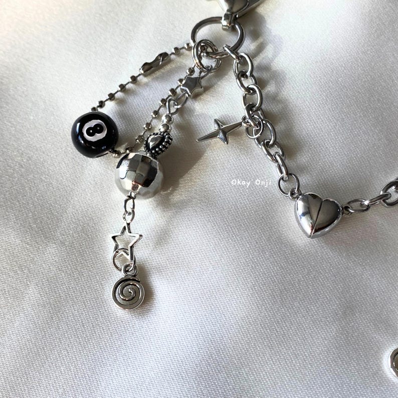 May include: Silver-coloured keychain with various charms. Charms include a black eight-ball, a disco ball, stars, a heart, and a spiral. The keychain is on a white fabric background.