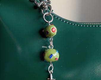 May include: A green and red beaded charm with a silver star charm attached to a silver chain. The charm is hanging from a green handbag.