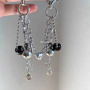 May include: Silver chain accessory with various charms. It includes disco ball beads, star and heart pendants, and black spherical beads. The accessory is attached to a silver clasp and ring.