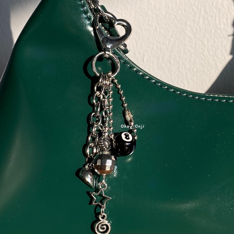 May include: Silver charm featuring a heart-shaped clasp and multiple dangling charms, including a star and a black 8-ball. The charm is attached to a silver chain. The text "Okay.Onji" is visible against a dark green background.