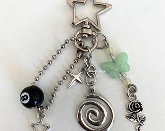 May include: Silver-toned keychain with a star-shaped clasp. Charms include a black 8-ball, a spiral, a rose, a butterfly, and multiple stars. The keychain has a ball chain and a small star charm.