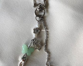 May include: A silver star-shaped keychain with a silver chain and charms. The charms include a green butterfly, a silver tooth, a silver flower, a silver star, and a silver swirl.