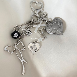 May include: A silver charm keychain with a variety of charms, including a heart locket, a cross, a star, a bow, a 8-ball, and a heart with an angel inside. The charms are attached to a silver chain with a silver lobster clasp.