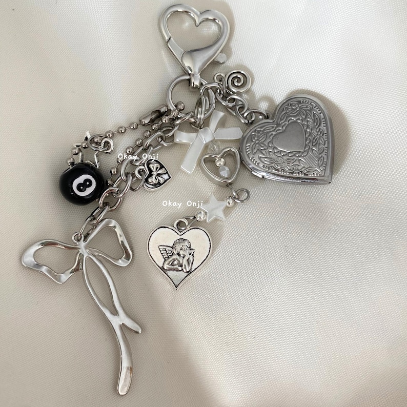 May include: A silver charm keychain with a variety of charms, including a heart locket, a cross, a star, a bow, a 8-ball, and a heart with an angel inside. The charms are attached to a silver chain with a silver lobster clasp.
