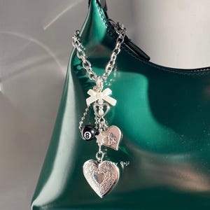 May include: A silver chain with charms attached to a green handbag. The charms include a white bow, a heart with a pearl, a black and white 8 ball, a star, a heart with angels, and a locket heart.