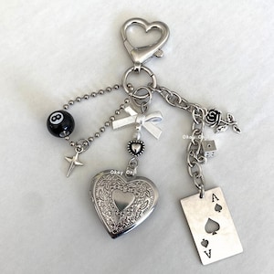 May include: Silver-toned keychain with various charms, including a heart-shaped locket, a black 8-ball, a star, a white bow, a rose, a die, and an Ace of Spades card. The keychain has a heart-shaped clasp.