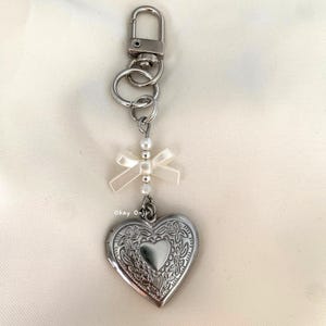 May include: A silver heart-shaped locket keychain with a white pearl and bow charm. The locket has a detailed floral design.