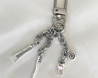 May include: Silver keyring with a toothbrush, toothpaste tube, and a tooth charm. The keyring also has a swirl charm and the text "Okay Oui" on it.