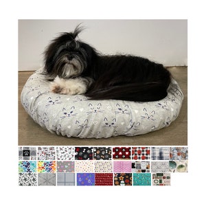 May include: A black and white dog is sleeping in a gray and white cat-themed donut bed. The bed has a white background with a repeating pattern of black cat faces and white hearts.