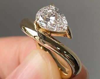 Pode incluir: Um anel de ouro com um diamante em forma de pêra e uma banda torcida. O anel está em um dedo.