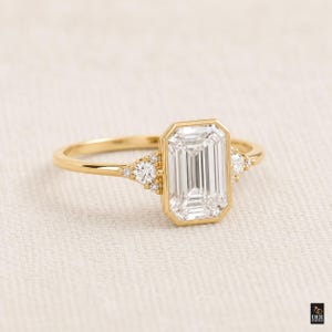 Könnte beinhalten: Ein goldener Ring mit einem Smaragd-Schliff-Diamanten. Der Mittelstein ist von einer goldenen Lünette umgeben, mit kleineren runden Diamanten auf jeder Seite. Das Band ist aus poliertem Gold.