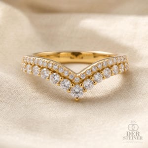 Könnte beinhalten: Ein goldener Ring in Chevron-Form mit zwei Reihen runder, im Brillantschliff geschliffener weißer Diamanten entlang des V-förmigen Bandes. Der Ring hat eine polierte Goldoberfläche und zeigt den Text DER STEINER EARTH AND LAB GROWN.