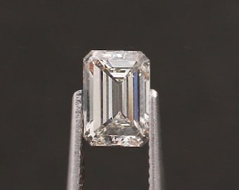 May include: An emerald-cut diamond held by metal tweezers. The rectangular diamond features a step-cut facet arrangement, displaying a clear, brilliant appearance. The gem is colourless and reflects light, set against a dark background.