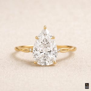 Könnte beinhalten: Ein goldener Verlobungsring mit einem birnenförmigen Diamanten. Das Band hat ein verdrehtes Design mit kleinen Akzentdiamanten. Der Ring ist vor einem neutralen Hintergrund platziert.