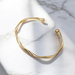 May include: A gold-toned twisted bangle bracelet with rounded ends. The bracelet is on a white marble surface.