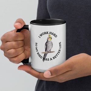 Puede incluir: Taza de café blanca con borde y asa negros. La taza tiene un círculo negro con el texto "Trabajo duro para que mi pájaro tenga una vida mejor" y una ilustración de acuarela de un cacatúa.