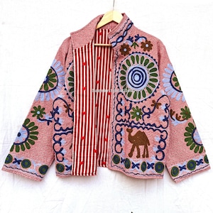 以下が含まれることがあります： 幾何学模様のピンク色の刺繍入りジャケット。青、緑、茶色で花柄とラクダのデザインが施されています。裏地は赤と白のストライプ。木製のハンガーにジャケットがかけられています。