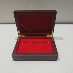 May include: A rectangular wooden display box with a dark maroon exterior finish and a vibrant red interior lining. The box features gold-coloured metal hinges and three rectangular recessed slots designed for holding items securely in place.