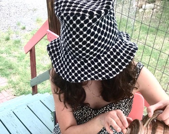 May include: A person wearing a black and white patterned bucket hat pours coffee from a brown ceramic teapot into a white mug with floral designs. The mug is sitting on a table with a blue and pink floral tablecloth.