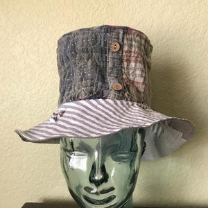 Puede incluir: Un sombrero de patchwork gris, marrón y blanco con un ala ancha y una banda a rayas. El sombrero está hecho de tela acolchada y tiene dos botones de madera en el lateral.