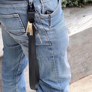 May include: A black key chain with a black strap and a set of keys attached. The keychain is hanging from the belt loops of a pair of blue jeans.