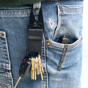 May include: A black key chain with a clip and a ring holding a set of keys and a black folding knife in a blue jean pocket.