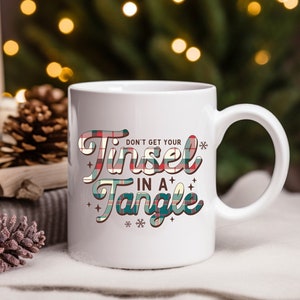 Puede incluir: Taza de cerámica blanca con el texto "DON'T GET YOUR TINSEL IN A TANGLE" en un diseño de cuadros. La taza tiene un asa curva y está decorada con copos de nieve y estrellas. Es un artículo festivo.
