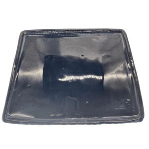 May include: A square, dark blue ceramic tray with a glossy finish. The tray has slightly rounded corners and a raised edge. The surface reflects light, highlighting subtle variations in the glaze. Suitable for serving or display.