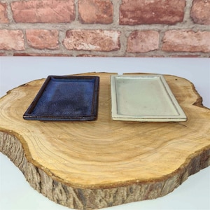 May include: Two rectangular ceramic dishes. One is a deep blue with a speckled glaze, the other is a light cream colour. Both are displayed on a natural wood slice, with a brick wall in the background.