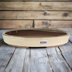 May include: A cream-coloured, oval bonsai pot with a brown interior. The pot has a low profile and a slightly raised rim. The number "0399" is printed on a white sticker on the side. The pot sits on a wooden surface.