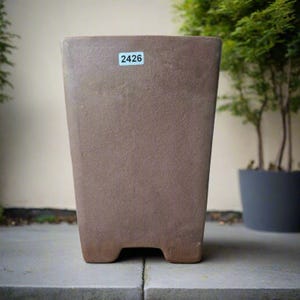 May include: A brown rectangular planter with a textured surface. The planter has a slightly tapered shape and sits on four small feet. A sticker with the number "2426" is affixed to the front. The background includes a glimpse of greenery.