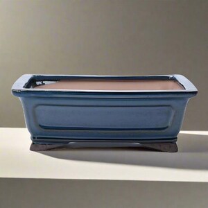 May include: A rectangular, blue ceramic bonsai pot with a brown interior. The pot has a slightly raised rim and sits on small, dark-coloured feet. The pot's exterior has a glossy finish.