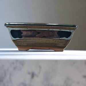 May include: Rectangular bonsai pot with a brown and black glazed finish. The pot has a dark rim and a light brown base with small feet. The sides feature a textured design with dark accents.