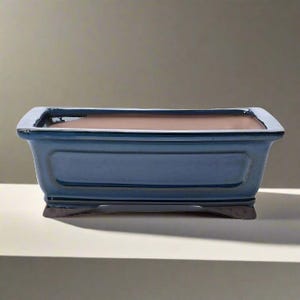 May include: A rectangular, blue ceramic bonsai pot with a brown interior. The pot has a slightly raised rim and sits on small, dark-coloured feet. The pot's exterior has a glossy finish.