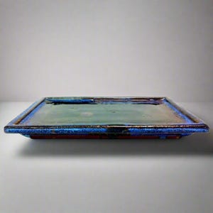 May include: A rectangular ceramic serving tray with a glossy finish. The tray has a deep blue border, a green centre, and hints of brown and red. The design is simple and elegant, suitable for serving food or as a decorative piece.