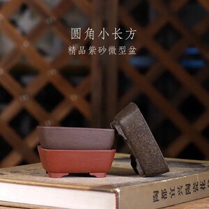 May include: Two rectangular terracotta bonsai pots, one brown and one red, with a book in the background. The text on the book is in Chinese. The text on the pots reads "Round Small Rectangle, Exquisite Clay Handmade Pot".