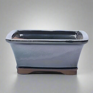 May include: A rectangular bonsai pot with a light blue-grey glaze. The rim is a shiny silver colour, and the base has a brown accent. The pot has a slightly flared shape, designed for planting small trees or plants.