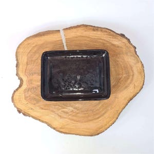 May include: A rectangular, dark brown soap dish with a glossy finish. The dish is placed on a round, light brown wooden slab with a natural edge. The background is white.