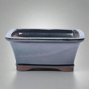 May include: A rectangular bonsai pot with a glossy, grey-blue finish. The rim is a shiny silver colour, and the base has a brown accent. The pot has a slightly flared shape and is designed for planting small trees or plants.