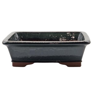 May include: A dark green rectangular bonsai pot with a glossy finish. The pot has a flared shape and rests on four small, brown feet. The interior has a lighter, textured appearance. This ceramic container is designed for bonsai trees.