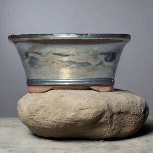 May include: A rectangular bonsai pot with a metallic blue glaze, resting on a large, rounded stone. The pot has a wide, flared shape with a dark rim and sits on small, rounded feet. The stone is a light brown colour.