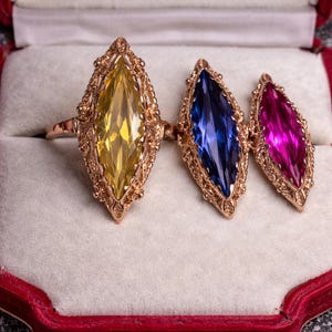 Peut inclure: Trois bagues ornées de pierres précieuses dans un écrin en velours. Les bagues présentent des pierres de forme marquise jaunes, bleues et magenta, serties dans des montures en filigrane dorées. L'écrin est rouge avec un intérieur blanc.